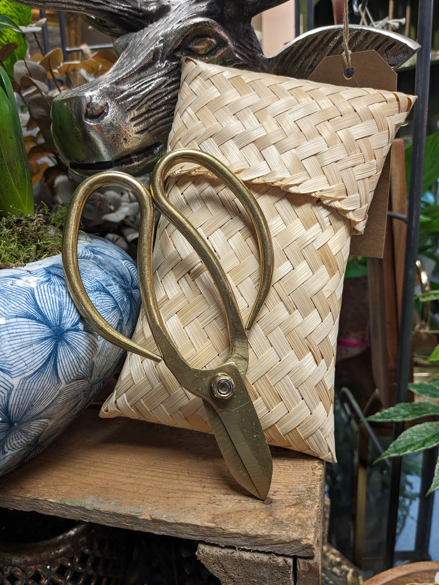 A pair of bronze-colored garden scissors with oversized looped handles, resting on a woven bamboo bag, set against a rustic garden backdrop.