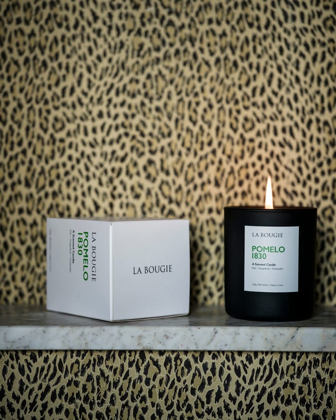 A black candle with a lit flame, next to a white box with 'LA BOUGIE' printed on it, against a leopard print background.