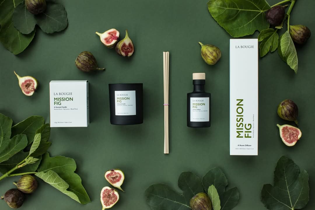 A room diffuser product laid out with fig leaves and figs on a green background, including a box, diffuser bottle, and reeds.