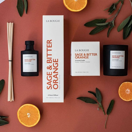 A product display featuring a black candle, a white box with packaging, a diffuser bottle, and decorative elements like orange slices and green leaves on a brown background.