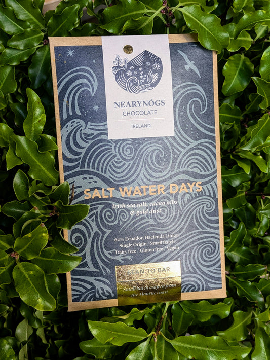 A chocolate bar named 'Salt Water Days' by Nearynog's Chocolate, placed on a bed of green leaves, with packaging that includes Irish sea salt, crushed cacao nibs, and gold dust.