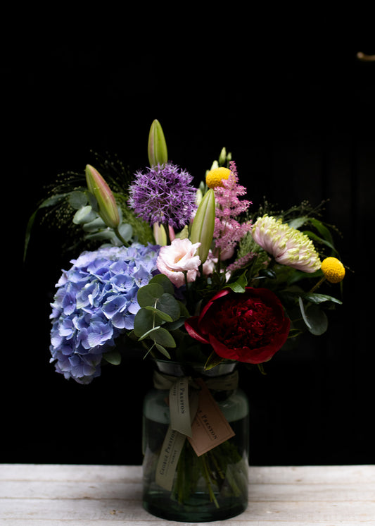 A bouquet of assorted flowers including purple, red, and yellow, arranged in a clear glass vase, placed on a table with a dark background.