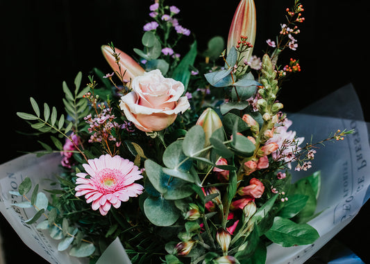 A hand-tied bouquet with a variety of flowers including roses and gerberas in shades of green and pink, wrapped in eco-friendly grey paper.