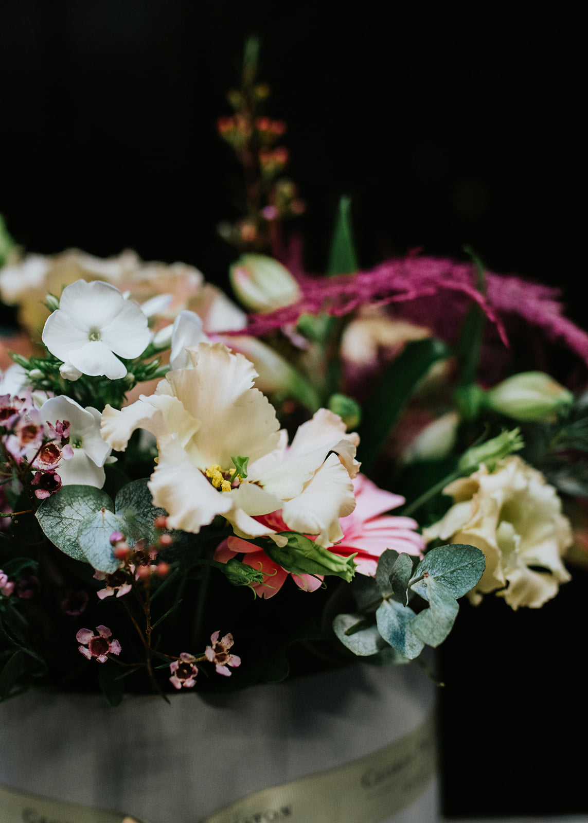 A floral arrangement featuring a variety of flowers including pink, white, and purple, presented in a clear hatbox.