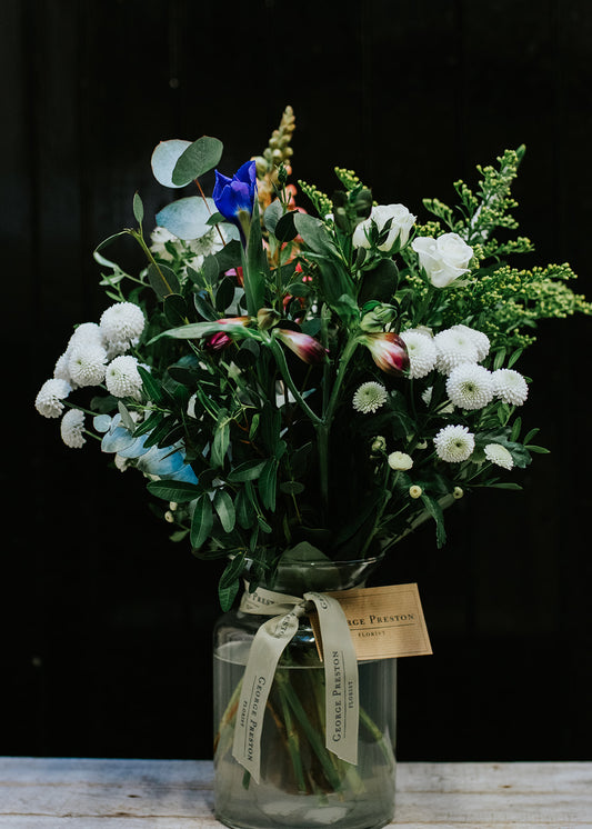 A glass vase containing a bouquet of flowers with green, blue, and white colors, tied at the bottom with a ribbon and a tag indicating the florist.